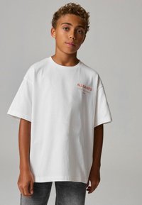 Bílé oversize tričko z bavlny s krátkými rukávy a kulatým výstřihem. Na přední straně se nachází červené logo "ALLSAINTS", stylizované s šedými džíny.