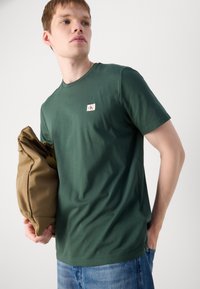 Vihreä puuvillapaita, jossa on pyöreä kaula-aukko ja lyhyet hihat, ja jossa on pieni logopatch rinnan kohdalla. Malli pitää käsissään vaaleaa laukku.