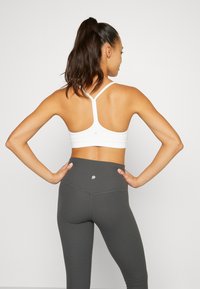 Balts sporta krūšturis ar sacensību aizmugures dizainu, apvienots ar augstās jostasvietas tumšā pelēkā krāsā ribotām legingām. Uz legingām ir vienkāršs logo elements.