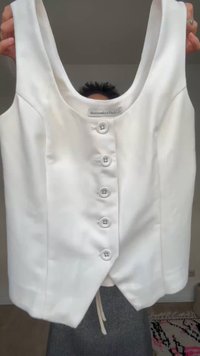 Biały bezrękawnik typu crop top zapinany na guziki, oznaczony marką Abercrombie & Fitch, trzymany w dłoniach na jednolitym tle.