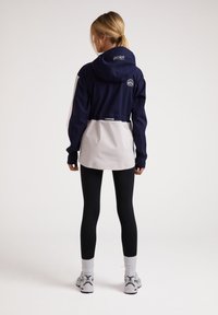 Chaqueta cortavientos en azul marino y blanca con capucha, que presenta una cintura ajustable, mangas largas y un logo en la parte posterior, combinada con leggins negros y zapatillas.