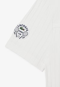 Vit tennis skjorta med en strukturerad, ribbad design. Den vänstra ärmen har en rund lapp med "LACOSTE TENNIS CLUB" och tennisgrafik.