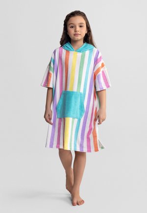 Młoda dziewczyna w kolorowym paskowanym ponczo z kapturem i przednią kieszenią, stojąca boso na jednolitym szarym tle.