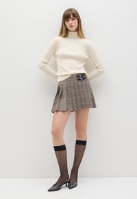 Maglione a collo alto beige a costine, gonna plissettata a quadretti con fibbie nere, collant trasparenti neri al ginocchio e tacchi a punta neri con pois bianchi.