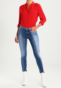 Chemise rouge boutonnée avec col, associée à un jean skinny bicolore bleu avec ourlet effiloché, et des bottines blanches à la cheville.