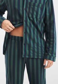 Pajama set s navy modrými a zelenými pruhy vyrobený z měkkého materiálu. Košile na knoflíky s náprsní kapsou a kalhoty s elastickým pasem.