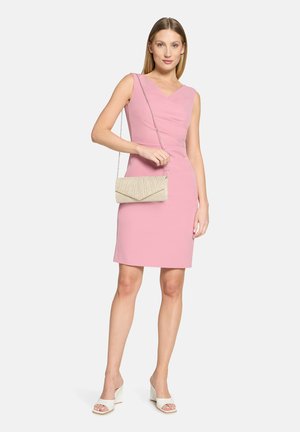 Robe rose sans manches avec une silhouette ajustée et un détail de tissu froncé. Le mannequin tient une pochette dorée avec des accents plissés. Sandales blanches à talons.