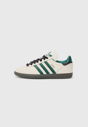 Baskets blanches Adidas Samba avec lacets noirs, bandes vertes et noires et semelle gris foncé sur fond uni.