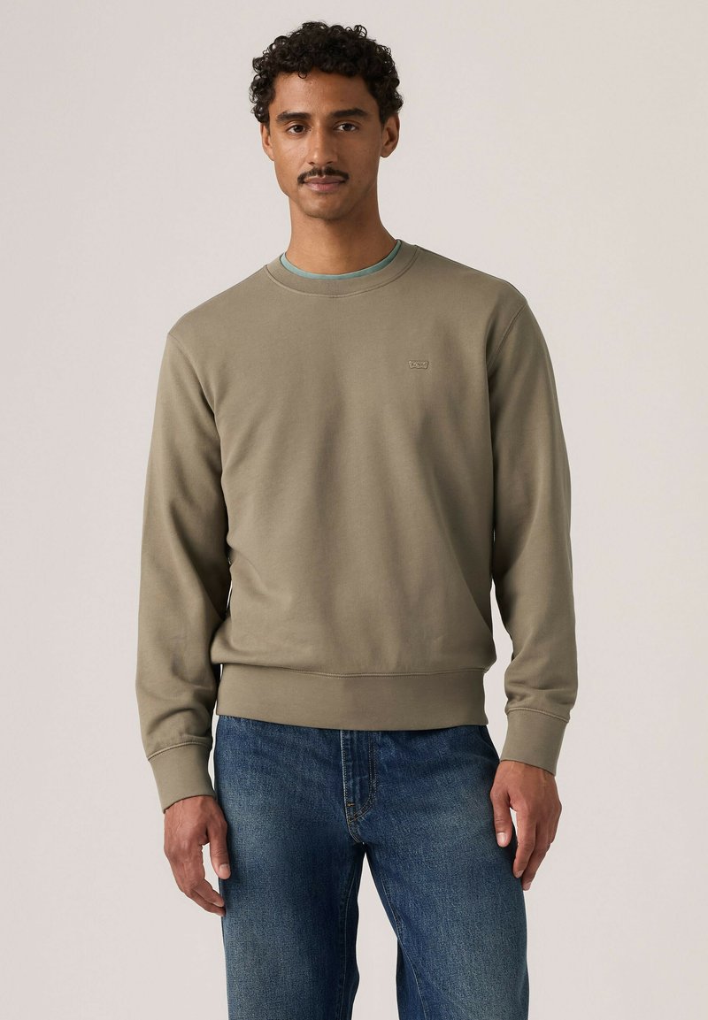 Mann mit kurzen lockigen Haaren und Schnurrbart, der einen hellbraunen Pullover und blaue Jeans trägt, steht vor einem einfarbigen Hintergrund.