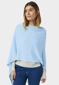 Kobieta z włosami do ramion, ubrana w jasnoniebieski ponczo na beżowym swetrze i niebieskich dżinsach, patrząca lekko na bok.