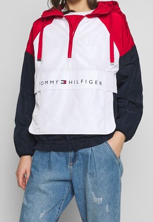 Persona che indossa un giubbotto antivento Tommy Hilfiger a blocchi di colore rosso, bianco e blu navy con logo e jeans blu chiaro leggermente consumati, con una mano in tasca.