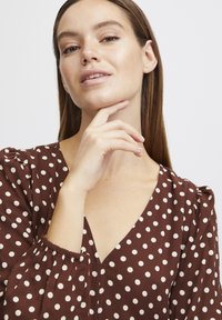 Blouse marron à pois avec manches bouffantes, décolleté en V profond et poignets froncés ; tissu lisse avec des pois crème contrastants et coupe décontractée.