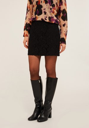 Falda mini de encaje negro con un patrón texturizado, combinada con botas negras de cuero con grabado de cocodrilo hasta la rodilla y un suéter multicolor.
