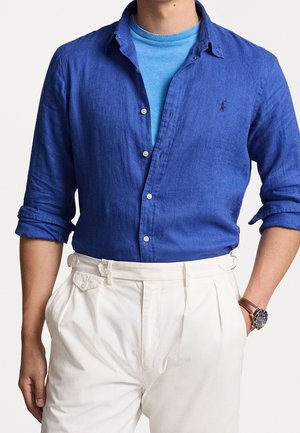 Homme portant une chemise bleue boutonnée par-dessus un t-shirt bleu clair, un pantalon blanc taille haute et une montre-bracelet marron avec un cadran noir.