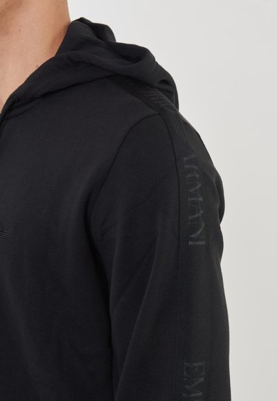 Felpa nera con zip, dotata di cappuccio, texture a coste e accenti logo tonali lungo le maniche. Design aderente con una silhouette classica.