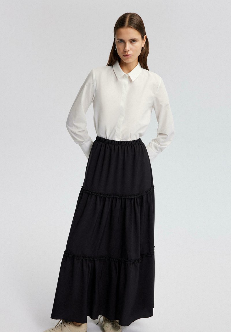 Zwarte maxi rok met gelaagd ontwerp, gerimpelde accenten en een elastische tailleband. Gepaard met een witte bloes met kraag.