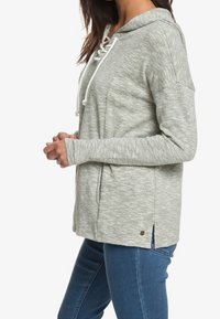 Sweat à capuche gris clair texturé avec un laçage à l'avant, des fentes latérales et un petit logo. Porté avec un jean bleu.