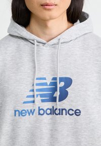 Svijetlosivi dukserica napravljena od mješavine pamuka s prednjim kengur džepom. Sadrži plavi New Balance logo i natpis u tamnoplavoj boji.