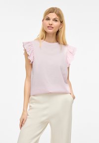 Helles rosa Kurzarmoberteil mit rüschierten Schulterakzenten; glatte Textur und lockere Passform. Getragen mit cremefarbenen weiten Hosen.