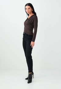 Vrouw met zwart haar die een donkerbruine langemouwen gerimpelde top draagt, zwarte strakke broek en zwarte enkellaarsjes met hoge hakken, staand opzij.
