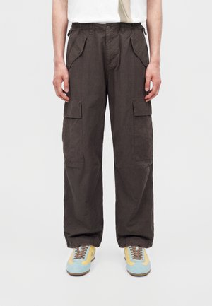 Persona che indossa pantaloni cargo marrone scuro con più tasche e scarpe da ginnastica azzurro chiaro e gialle su uno sfondo uniforme.