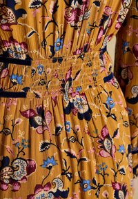 Blumenkleid aus senfgelbem Stoff mit rosa, blauen und marineblauen Mustern. Strukturierte Oberfläche mit gerafftem Taillenbund und Rüschendetails.