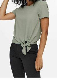 Top khaki a maniche corte con scollatura a girocollo, dotato di un nodo all'orlo anteriore e una piccola tasca, abbinato a pantaloni neri aderenti.