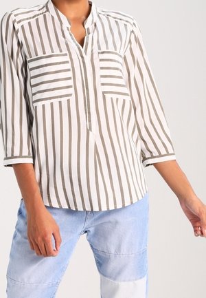 Vrouw draagt een witte blouse met verticale beige strepen en twee gestreepte borstzakken, gecombineerd met lichtblauwe patchwork spijkerbroek.