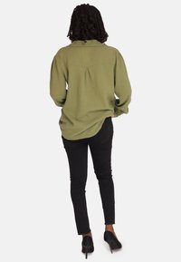 Camisa de manga larga verde oliva con tejido texturizado, corte relajado, pliegue trasero y cuello doblado, combinada con pantalones negros ajustados y tacones negros.