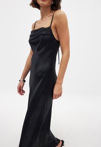 Musta satiini mekko, jossa on kaareva kaula-aukko ja ohuet säädettävät olkaimet. Tiukka siluetti, lattiaa hipova pituus, sileä tekstuuri ja minimaliset suunnitteluyksityiskohdat.