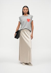 Pulover gri, fără mâneci, cu un grafic în formă de inimă roșie, asociat cu o fustă bej, lungă până la podea, din satin. Modelul ține o geantă de mână neagră.