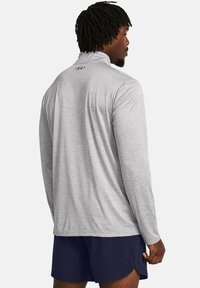 Graues langärmliges Sportshirt mit hohem Kragen und strukturierter Stoffqualität, mit einem kleinen Logo auf dem Rücken. Kombiniert mit navyblauen Shorts.