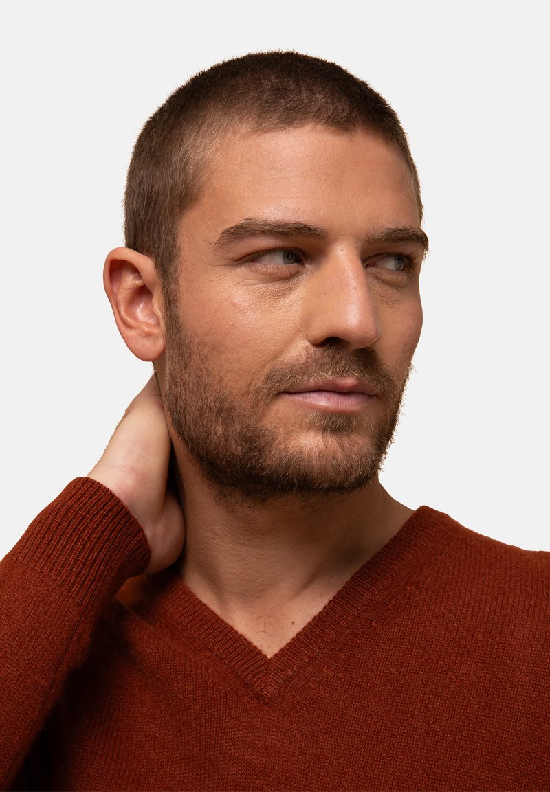 Sweter w serek w kolorze spalanej pomarańczy, wykonany z miękkiej tkaniny. Posiada ściągacze na rękawach i fakturowane wykończenie, noszony przez osobę z krótkimi włosami.