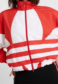 Röd och vit vindjacka med dragkedja, djärva ränder, hög krage samt elastisk nederkant och ärmslut. Lätt, texturerad tyg.