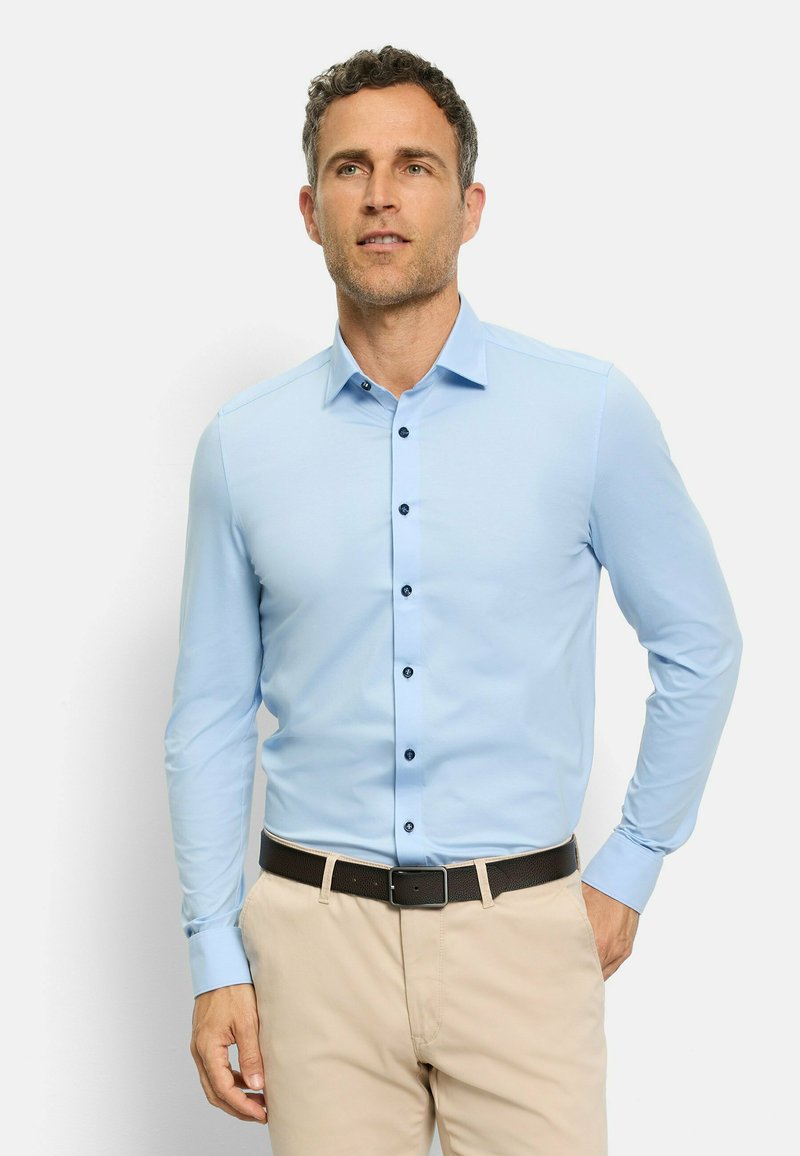 Chemise à manches longues bleu clair avec une coupe ajustée, boutons noirs et col contrastant. Associée à un pantalon beige et une ceinture foncée.