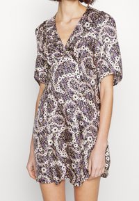 Femme portant une robe courte à fleurs avec des manches bouffantes et un décolleté en V aux couleurs violettes, blanches et jaunes.