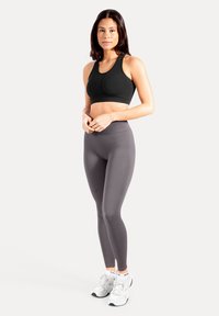 Brassière de sport noire à encolure dégagée, associée à des leggings gris ajustés et taille haute. Mannequin portant des chaussures de sport blanches. Texture lisse.