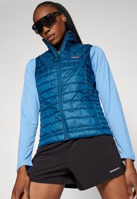 Gilet rembourré bleu avec un col haut, texture matelassée, fermeture éclair à l'avant, porté sur une chemise à manches longues bleu clair et un short noir.