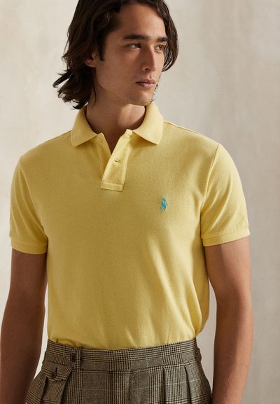 Jaunas vyras su vidutinio ilgio plaukais, dėvintis geltoną polo marškinėlį su mažyčiu mėlynu siuvinėtu logotipu ir rudomis languotomis kelnėmis.