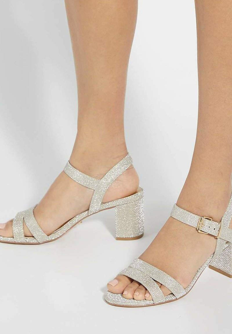 Sandales à talons hauts argentées avec une finition texturée et pailletée. Comprend des brides croisées, une boucle à la cheville et un design de talon bloc.