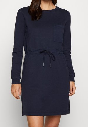 Kobieta ubrana w granatową, długorękawową sukienkę casual z kieszenią na piersi i regulowanym ściągaczem w talii, stoi na tle jednolitego tła.