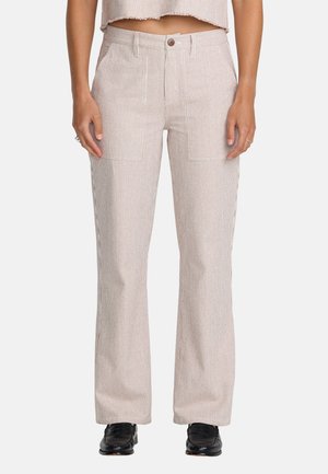 Kobieta w beżowych, szerokich spodniach w prążki z wysokim stanem, z przednimi naszywanymi kieszeniami i czarnymi butami, stoi na jednolitym tle.