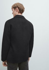 Czarny wełniany płaszcz o strukturalnym kroju, z klasycznym kołnierzem i długimi rękawami. Tył gładki, z lekko zaokrąglonym dołem i bez widocznych zapięć.