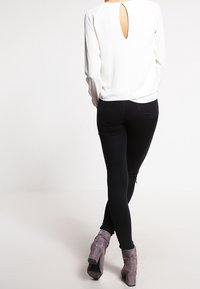 Weißes Langarmshirt mit Schlüsselloch-Detail auf dem Rücken, kombiniert mit schwarzen Slim-Fit-Jeans und grauen Stiefeletten. Glatter Stoff, figurbetontes Design und schlanke Silhouette.