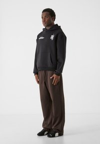 Sort hættejakke med frontlomme, udstyret med et logo på brystet. Sammen med løse brune sweatpants og sorte sportssko.
