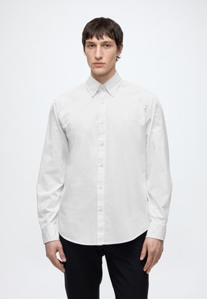 Homme portant une chemise blanche unie à manches longues boutonnée et un pantalon noir, debout devant un fond blanc.