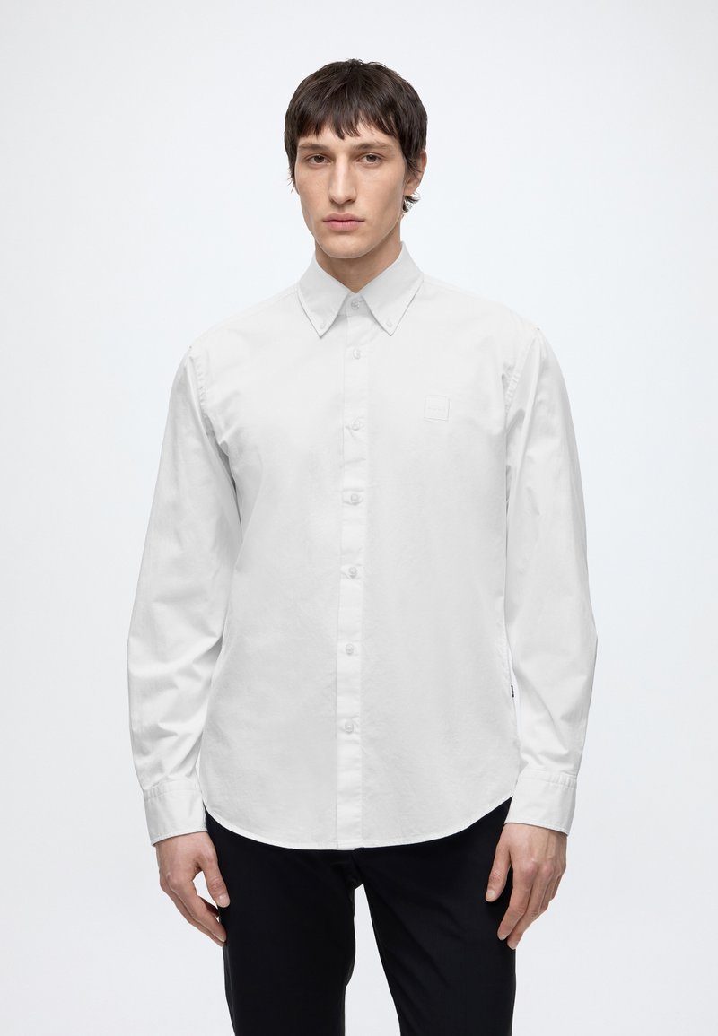Homme portant une chemise blanche unie à manches longues boutonnée et un pantalon noir, debout devant un fond blanc.