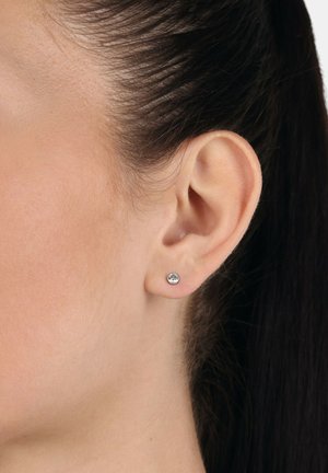 Silberne Steckohrringe mit rundem, klarem Edelstein. Glatte Metalloberfläche. Am Ohrläppchen getragen, mit seitlich gekämmtem Haar. Schlichtes Design.
