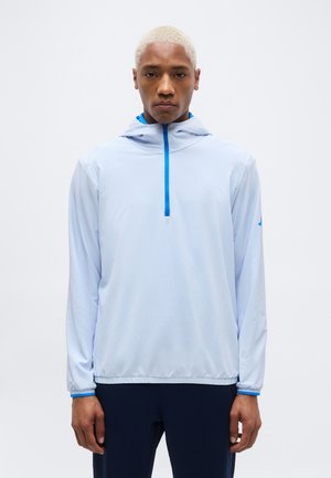 Junger Mann mit kurzen blonden Haaren, der einen hellblauen Kapuzenpullover mit einem blauen Reißverschluss trägt und vor einem schlichten weißen Hintergrund steht.