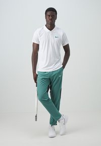 Witte polo met groen Nike-logo, gecombineerd met teal trainingsbroek met zwarte zijpanelen. Model houdt een tennisracket vast en draagt witte sneakers.